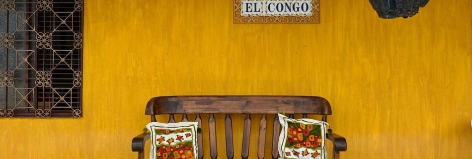 fachada de una casa amarilla con azulejos que dicen "El Congo" y una maceta colgando con flores moradas. Al frente hay una banca de madera con dos cojines