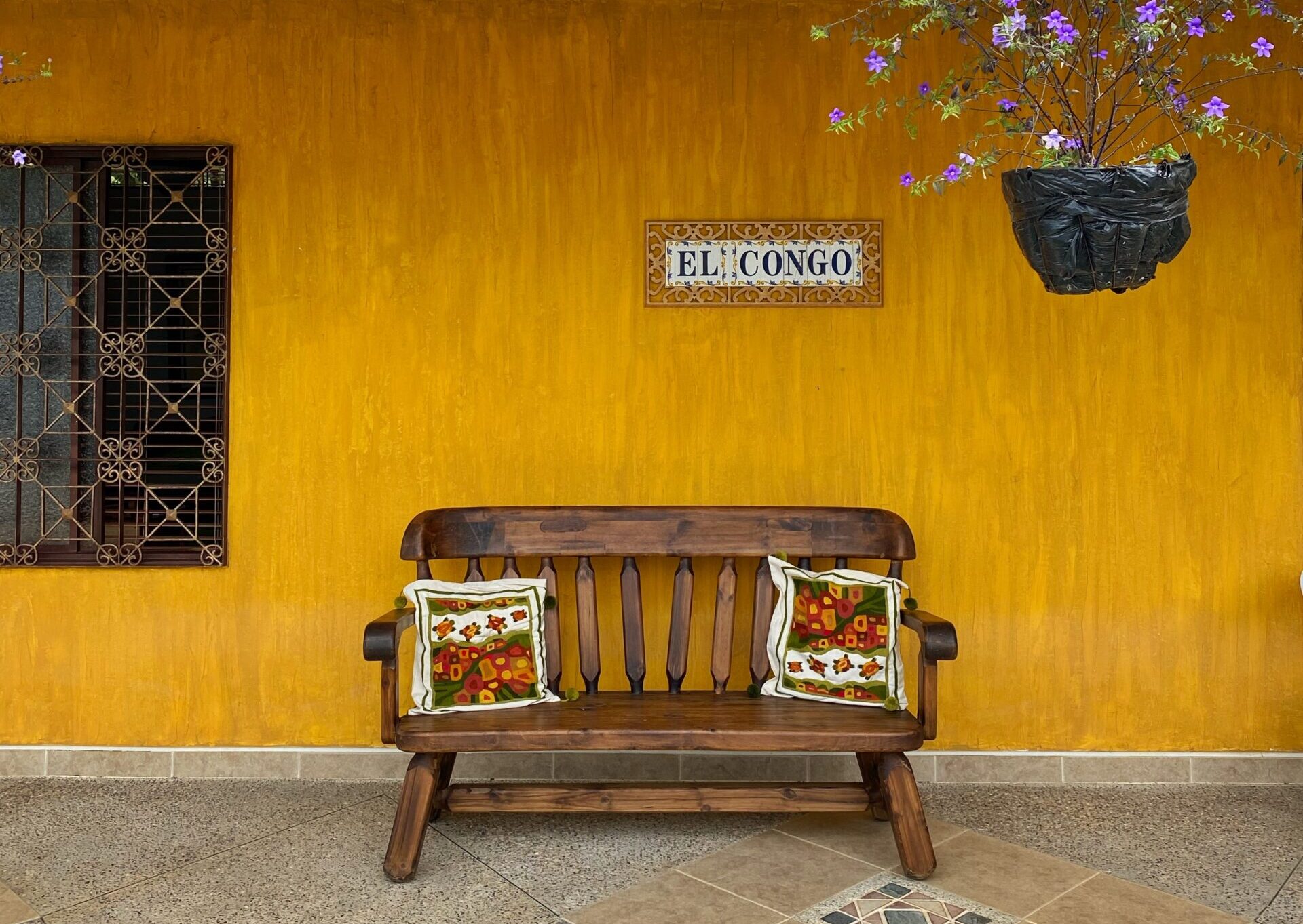 fachada de una casa amarilla con azulejos que dicen "El Congo" y una maceta colgando con flores moradas. Al frente hay una banca de madera con dos cojines