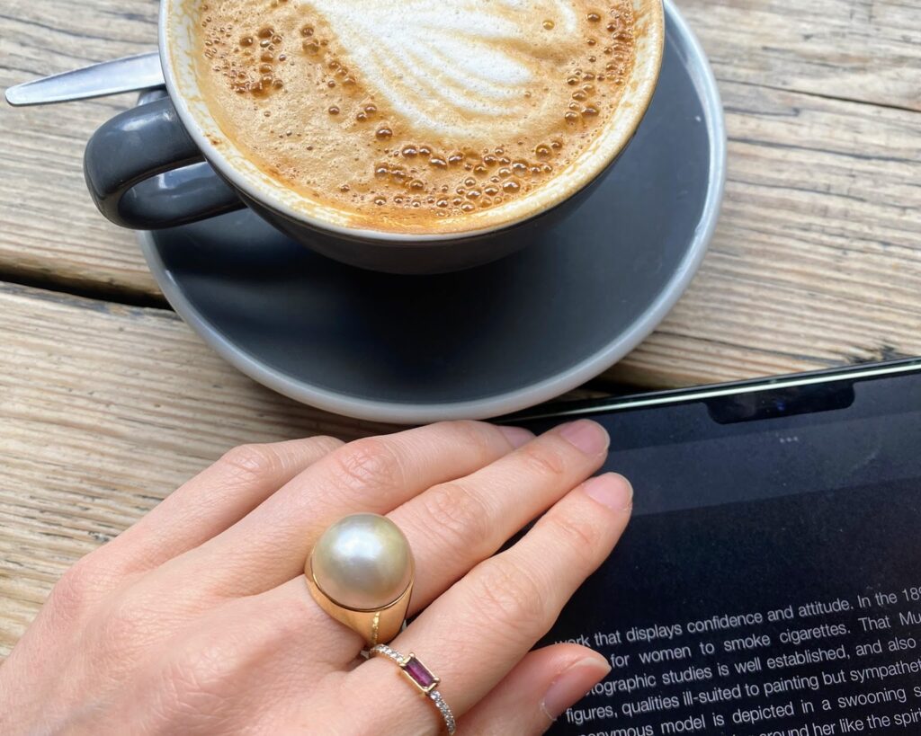Table set detail, featuring a cup of cappuccino, Laura's hand with a large pearl ring and a smaller ring with a rectangular ruby and small diamonds, and a tablet screen featuring some text on fashion film.
